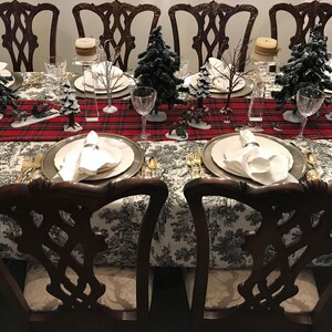 May include: A formal dining table setting with a red and black plaid tablecloth, white napkins, and silver flatware.  The table is decorated with small artificial pine trees and candles.