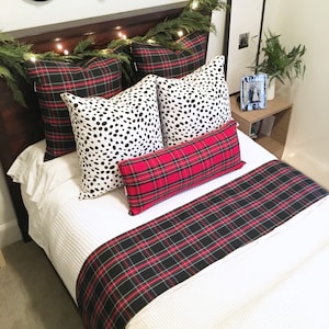 May include: A white bed with a red and black plaid bed runner and throw pillows. The pillows are a mix of black and white polka dot and red and black plaid patterns. The bed is decorated with a garland of greenery and lights.