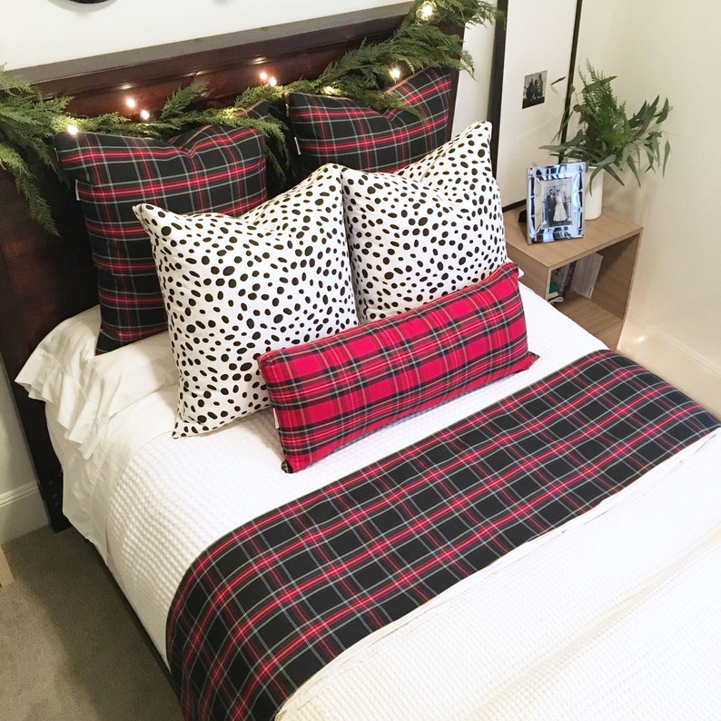 May include: A white bed with a red and black plaid bed runner and throw pillows. The pillows are a mix of black and white polka dot and red and black plaid patterns. The bed is decorated with a garland of greenery and lights.