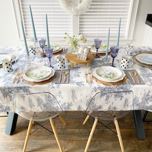 May include: A table setting for a spring or Easter dinner party. The table is covered in a white tablecloth with a blue floral pattern. There are blue and white polka dot mugs, blue glass goblets, and white plates with a floral design. There are also blue candles and a wooden tray with a small white basket filled with white eggs.