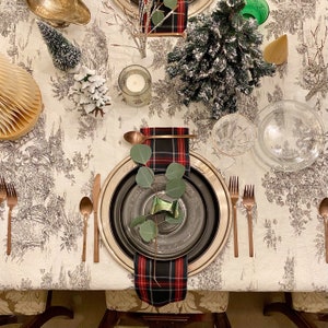 May include: A festive table setting featuring a black and red tartan napkin, a black and silver plate, and a sprig of eucalyptus. The table is adorned with a white tablecloth with a floral design.