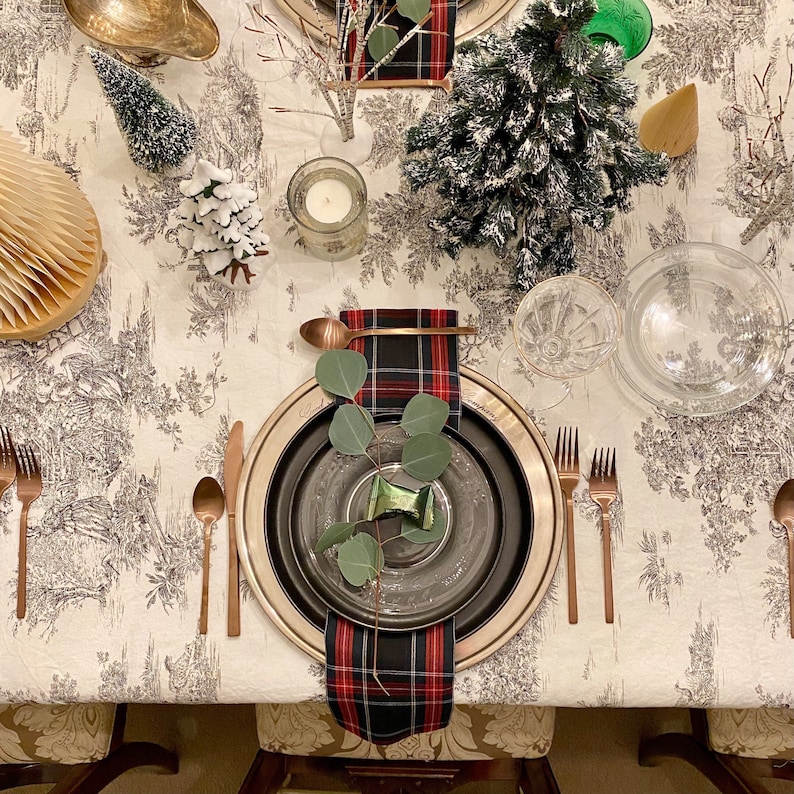 May include: A festive table setting featuring a black and red tartan napkin, a black and silver plate, and a sprig of eucalyptus. The table is adorned with a white tablecloth with a floral design.