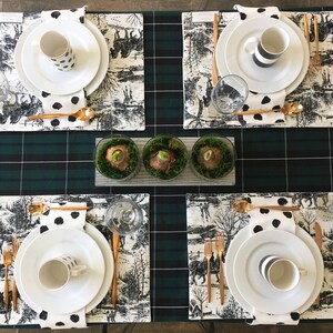 May include: A table setting with four place settings, each with a white plate, a black and white patterned mug, a gold fork and knife, and a black and white patterned napkin. The table is covered in a green and black plaid tablecloth and has a row of three small glass vases with green moss and white bulbs.