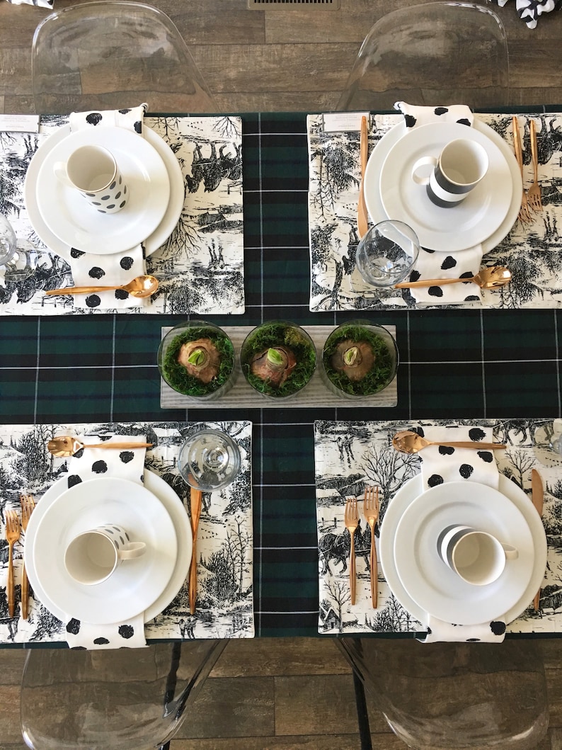May include: A table setting with four place settings, each with a white plate, a black and white patterned mug, a gold fork and knife, and a black and white patterned napkin. The table is covered in a green and black plaid tablecloth and has a row of three small glass vases with green moss and white bulbs.