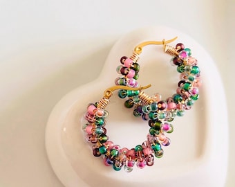 May include: Gold-coloured hoop earrings, each decorated with a multitude of small, colourful beads in shades of pink, green, purple, and clear. The earrings are displayed on a white, heart-shaped dish.