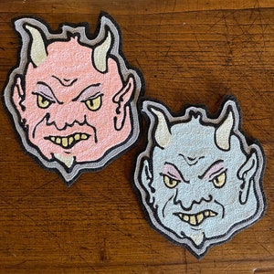 May include: Two embroidered devil face patches. One is light pink with yellow eyes, cream horns, and a gray outline. The other is light blue with yellow eyes, cream horns, and a gray outline. Both have a black border.