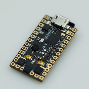 May include: A black circuit board with gold-colored connectors and a micro USB port. The board has several components, including a chip, a resistor, and a capacitor. The text "INJ" is printed on the board.