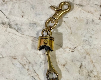 May include: A gold-toned key chain featuring a piston-shaped charm with a black key attached. The key chain has a gold-toned clasp.