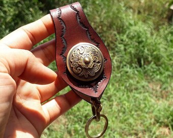 May include: A brown leather keychain with a decorative brass button. The keychain has a carved design along the edges and a brass key ring. The button features an ornate floral pattern.