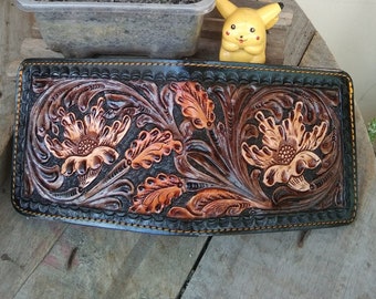 Peut inclure: Un portefeuille en cuir fait à la main avec un motif floral détaillé. Le portefeuille présente des fleurs et des feuilles en relief dans des tons de marron et de cuivre sur un fond sombre. Les bords sont cousus avec du fil jaune.