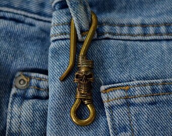 May include: A brass skull keychain with a hook and a loop. The keychain is attached to a pair of blue jeans.