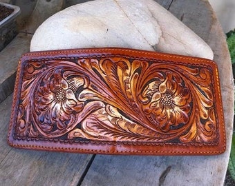 May include: A brown leather wallet with intricate floral tooling. The wallet has a stitched edge and a flap closure.