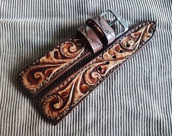 Könnte beinhalten: Ein braunes Leder-Uhrenarmband mit aufwendiger Blumenschnitzerei. Das Armband hat eine silberne Schnalle und wird auf einem gestreiften Stoffhintergrund gezeigt. Das Leder hat einen satten, warmen Farbton.