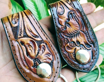 May include: Two brown leather keychains with intricate floral designs and silver metal rings. The keychains have a brown leather strap with a silver metal button.