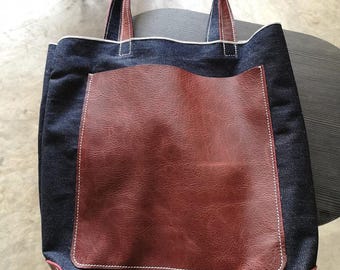 May include: A dark blue denim tote bag featuring a reddish-brown leather pocket. The bag has matching leather handles and trim, with white stitching. The bag is designed for carrying items. The bag's measurements are in centimeters.