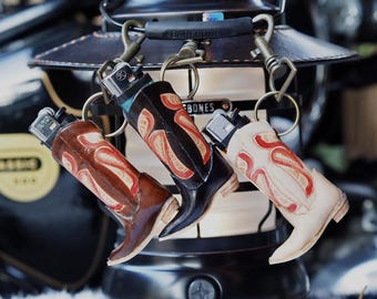 May include: Three leather boot-shaped keychains with metal rings and lighters inside. The boots are decorated with red and white patterns. The keychains are hanging from a leather strap with the word "BONES" on it.