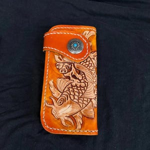 Könnte beinhalten: Eine orangefarbene Ledergeldbörse mit einem geprägten Koi-Karpfen-Design. Die Geldbörse hat einen Klappenverschluss mit einem silbernen Knopf und einem türkisfarbenen Mittelteil. Die Kanten sind mit weißem Faden genäht, und das Design ist aufwendig und handgefertigt.