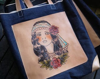 May include: A denim tote bag with leather accents and a hand-tooled leather patch featuring a woman with a floral design.
