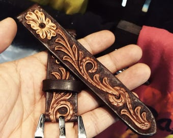 Könnte beinhalten: Ein braunes Leder-Uhrenarmband mit floralem und Ranken-Design. Das Armband hat eine geschnitzte Blume und dekorative Muster. Das Uhrenarmband hat eine silberne Schnalle.