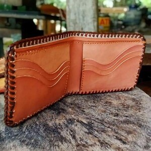 Puede incluir: Una cartera de cuero marr&oacute;n con un borde cosido y m&uacute;ltiples ranuras para tarjetas. La cartera est&aacute; abierta y el interior es visible.
