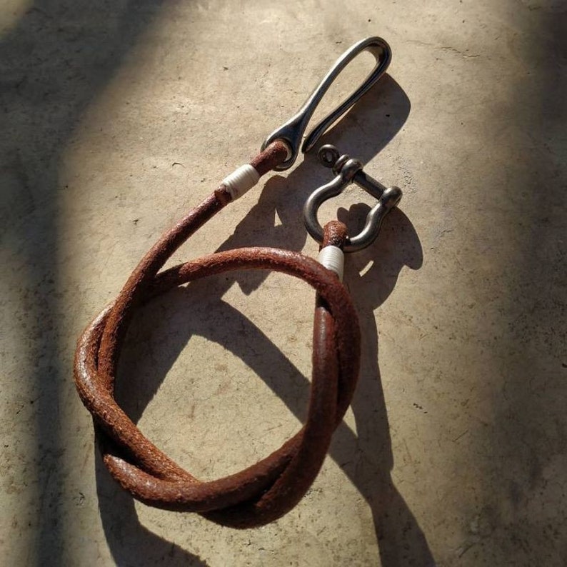 May include: A brown leather key chain with a silver hook and a silver D-ring. The leather is twisted and knotted.