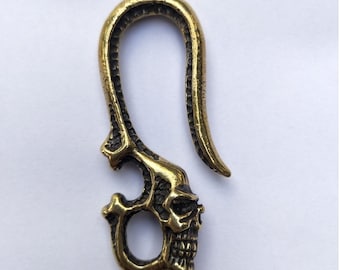 May include: A brass-colored, hook-shaped pendant featuring a skull design. The pendant has a curved top and a detailed skull at the bottom, with an open eye socket and jaw. The metal has an aged, antique appearance.