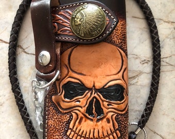 Könnte beinhalten: Eine braune Ledergeldbörse mit einem Totenkopfdesign und einem Metallconcho mit einem Bild eines indianischen Häuptlings. Die Geldbörse hat eine Kette und einen geflochtenen Lederriemen.