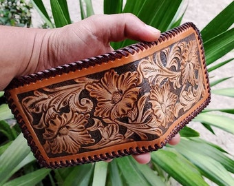 Puede incluir: Una cartera de cuero marrón con intrincados detalles florales. La cartera tiene un borde cosido y un fondo marrón oscuro.