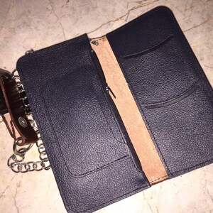 Puede incluir: Una cartera de cuero negro con una cadena adjunta. La cartera tiene varios bolsillos y un compartimento con cremallera. El interior est&aacute; forrado con cuero marr&oacute;n.