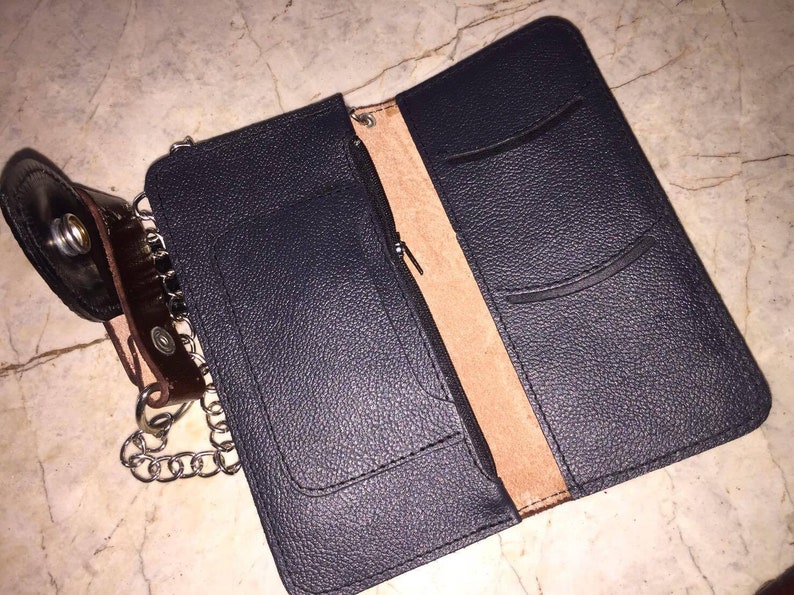 Puede incluir: Una cartera de cuero negro con una cadena adjunta. La cartera tiene varios bolsillos y un compartimento con cremallera. El interior est&aacute; forrado con cuero marr&oacute;n.