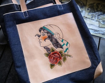 May include: A blue denim tote bag with a leather patch featuring a hand-tooled design of a woman with a rose. The woman is wearing a white hat with a green and blue stripe.