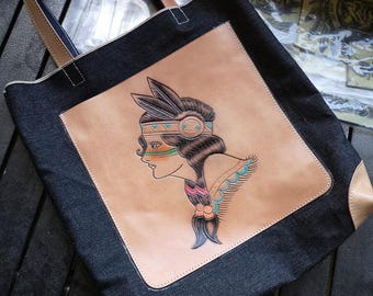 May include: A denim tote bag with a leather patch featuring a hand-drawn illustration of a Native American woman with feathers in her hair. The illustration is in a traditional tattoo style with colourful accents.