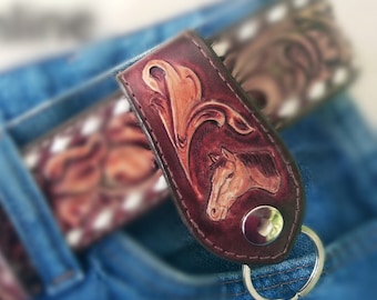 May include: A brown leather keychain with a tooled design of a horse's head and floral accents. The keychain has a silver key ring and a metal button.