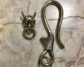 May include: A brass key chain with a detailed demon face charm and a coiled hook. The hook has a small ring at the bottom.