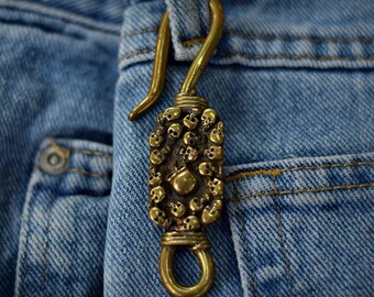 May include: A brass-colored keychain hook with a skull design, attached to a blue denim pocket. The hook is curved and has a rectangular pendant covered in small skulls. The keychain is attached to a denim pocket.