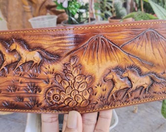 May include: A brown leather wallet with a tooled design of two horses running through a mountain landscape. The wallet has a stitched edge and a brown leather interior.