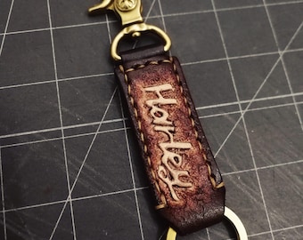 May include: A brown leather keychain with a brass hook and ring. The leather is embossed with the word "Harley" in a script font.