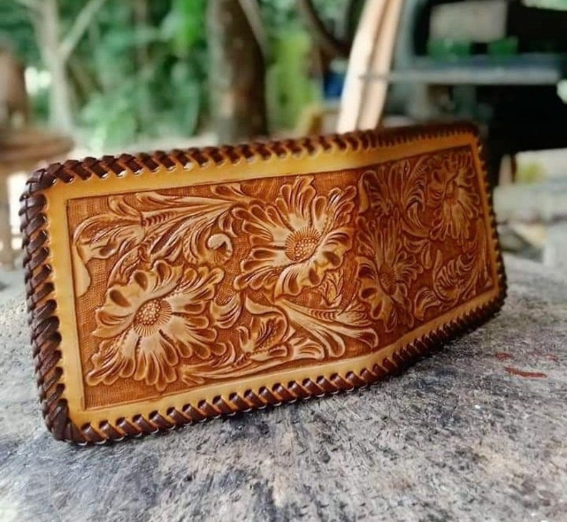 Puede incluir: Una cartera de cuero marr&oacute;n con un dise&ntilde;o floral tallado. La cartera tiene un borde trenzado y est&aacute; cerrada.