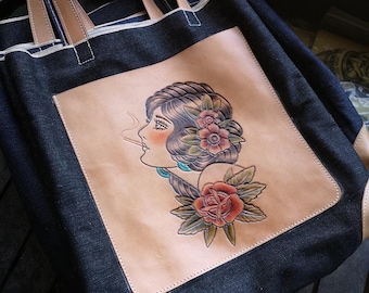 May include: A denim tote bag with leather straps and a leather patch featuring a hand-tooled design of a woman with flowers in her hair.