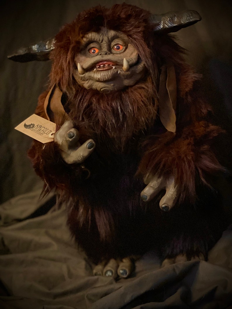 K&ouml;nnte beinhalten: Eine detaillierte, handgefertigte Pl&uuml;schfigur einer Kreatur mit braunem Fell, H&ouml;rnern und gro&szlig;en Ohren. Die Figur hat ein detailliertes Gesicht mit orangefarbenen Augen und einem Etikett mit der Aufschrift "KREALO ARTWORKS".