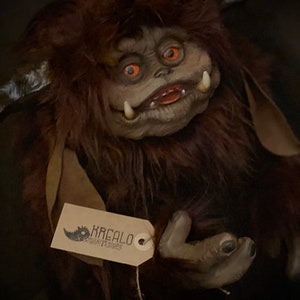 K&ouml;nnte beinhalten: Eine handgefertigte Kreaturenpuppe mit braunem Fell, gro&szlig;en Augen und einem grinsenden Mund. Die Puppe hat ein Etikett mit der Aufschrift "KREALO CREATURES". Das Gesicht der Puppe ist hellgrau und sie hat H&ouml;rner.