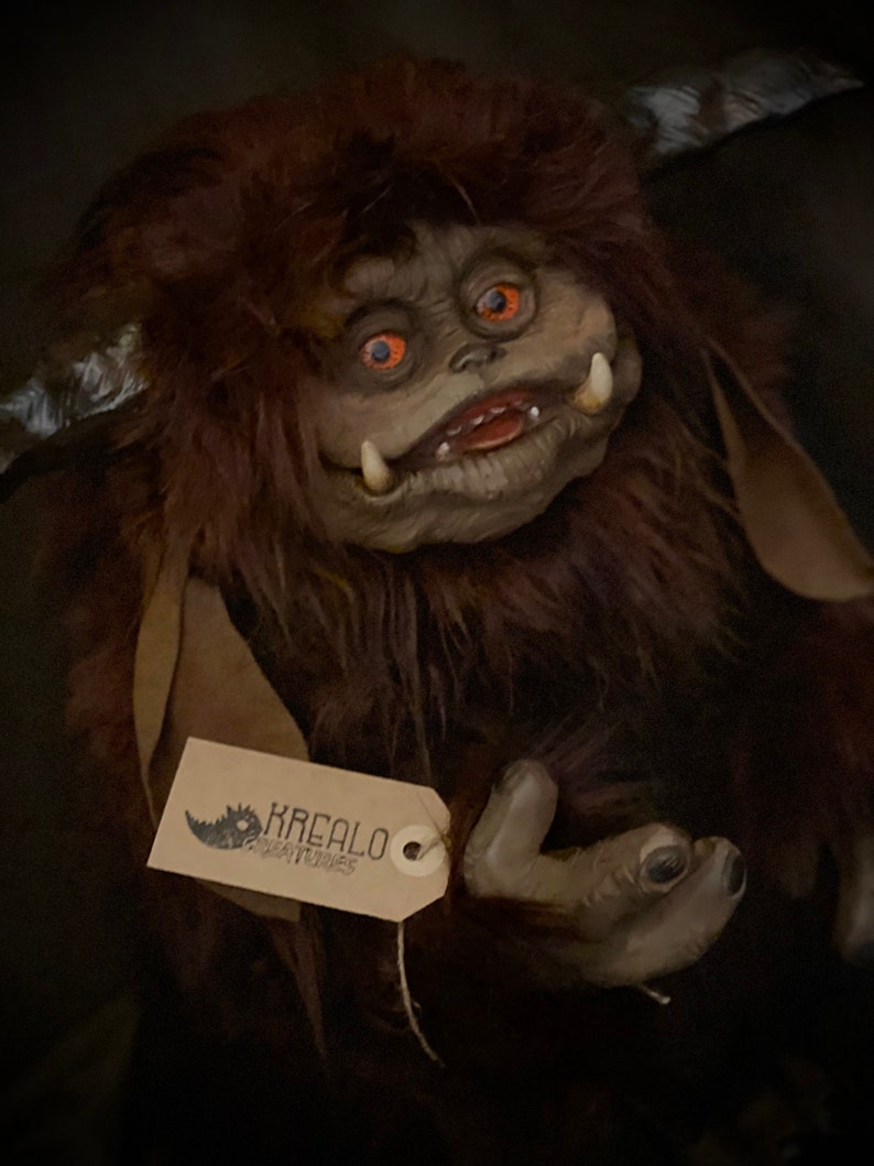 K&ouml;nnte beinhalten: Eine handgefertigte Kreaturenpuppe mit braunem Fell, gro&szlig;en Augen und einem grinsenden Mund. Die Puppe hat ein Etikett mit der Aufschrift "KREALO CREATURES". Das Gesicht der Puppe ist hellgrau und sie hat H&ouml;rner.