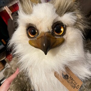 Puede incluir: Un b&uacute;ho de peluche blanco y marr&oacute;n con grandes ojos dorados. El b&uacute;ho tiene un pico marr&oacute;n y una etiqueta que dice "Krealoo Creatures".