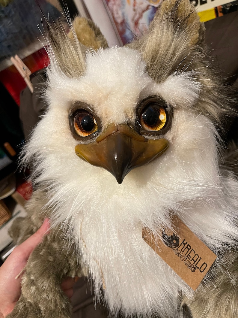 Puede incluir: Un b&uacute;ho de peluche blanco y marr&oacute;n con grandes ojos dorados. El b&uacute;ho tiene un pico marr&oacute;n y una etiqueta que dice "Krealoo Creatures".
