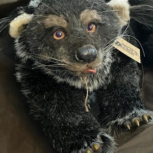 May include: A plush, handcrafted bear with a detailed, realistic design. The bear features a mix of black, gray, and tan fur, with expressive amber eyes and a small pink tongue. It has a tag attached, and its paws have detailed claws.