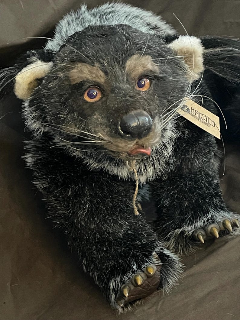 May include: A plush, handcrafted bear with a detailed, realistic design. The bear features a mix of black, gray, and tan fur, with expressive amber eyes and a small pink tongue. It has a tag attached, and its paws have detailed claws.