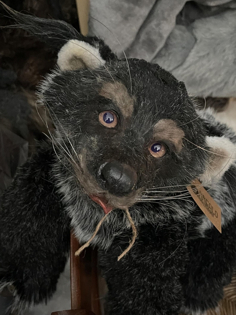 May include: A handmade plush toy in the shape of a raccoon. The toy has black and gray fur, with white accents on the ears and face. It has a brown tag attached. The raccoon has a realistic appearance with detailed features.