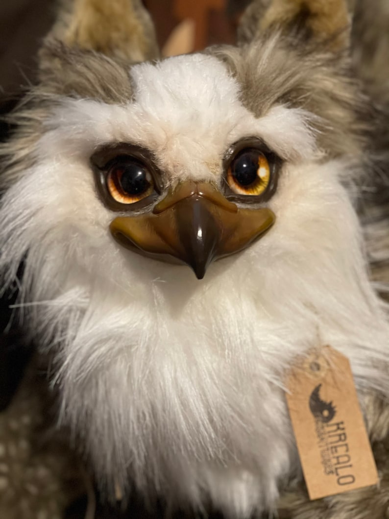Puede incluir: Primer plano de un juguete de peluche con la cara blanca, ojos marrones y un pico marr&oacute;n. El juguete tiene un patr&oacute;n de pelaje blanco y marr&oacute;n. El juguete tiene una etiqueta que dice "KreaLo Design & Toys".