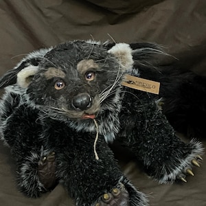 May include: A handmade plush toy of a black and gray animal with a long tail, possibly a bear or a raccoon. The toy has detailed features, including a realistic face, paws, and a tag that reads "MACALO". The plush is lying down on a brown surface.