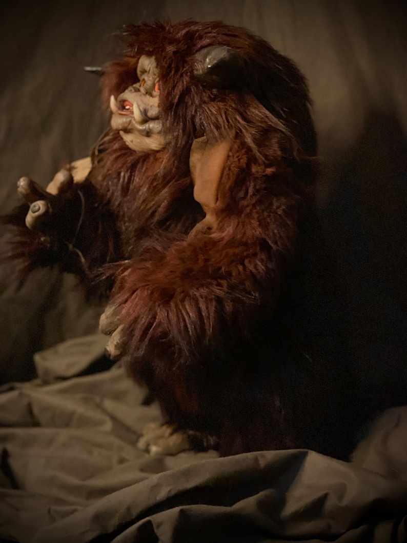K&ouml;nnte beinhalten: Eine braune, pelzige Monsterfigur mit detailliertem Gesicht, H&ouml;rnern und roten Augen. Die Kreatur hat einen bedrohlichen Ausdruck und steht aufrecht. Die Figur besteht aus einem weichen, pl&uuml;schigen Material.